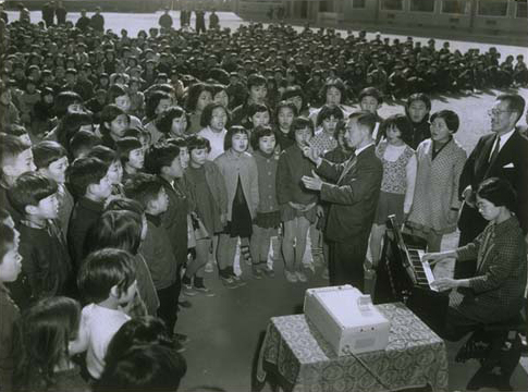 画像：1960（昭和35）年2月22日、名古屋市牧野小学校へリコーシンクロファックスを寄贈。児童たちが校歌を歌う姿を笑顔で見守る市村清（右端）